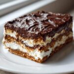 A close-up of a rich, layered chocolate eclair cake slice showing graham cracker layers, white cream filling, and thick chocolate frosting.