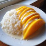 A plate of delicious mango sticky rice, featuring bright yellow mango slices over sweet rice drizzled with coconut cream.