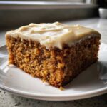 A close-up of a moist slice of applesauce cake topped with thick, creamy frosting on a white plate.