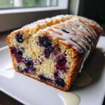Close-up of a slice of moist blueberry bread topped with a thick vanilla glaze drizzle.
