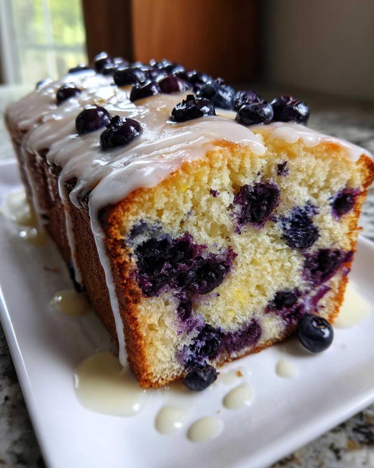 A slice of moist blueberry bread topped with vanilla glaze and fresh blueberries on a white plate.