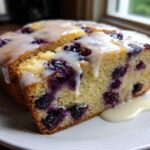 A close-up of a moist slice of blueberry bread topped with a thick, dripping vanilla glaze.