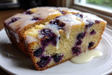 A close-up of a moist slice of blueberry bread topped with a thick, dripping vanilla glaze.