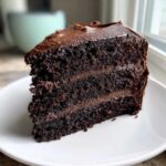 A rich, moist slice of dark chocolate cake recipe layered with thick chocolate frosting on a white plate.