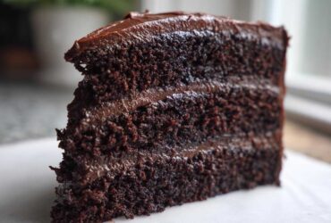 Close-up of a moist, dark slice of three-layer chocolate cake recipe with rich frosting.