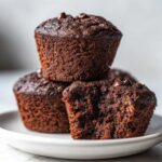 A stack of three rich, dark chocolate zucchini muffins, one broken open showing the moist texture and green zucchini flecks.