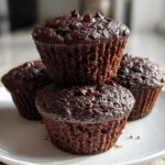 A stack of three rich, dark chocolate zucchini muffins topped with chocolate chips, resting on a white plate.