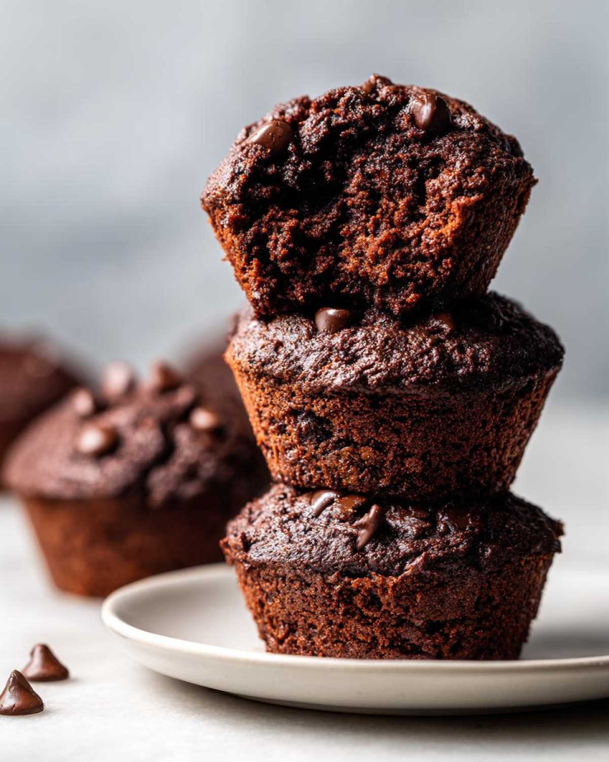A stack of three moist chocolate zucchini muffins topped with chocolate chips, sitting on a small white plate.