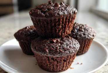 A stack of three rich, dark chocolate zucchini muffins topped with chocolate chips, resting on a white plate.