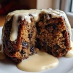Close-up of a rich, dark plum pudding with a piece cut out, generously topped with vanilla custard sauce.