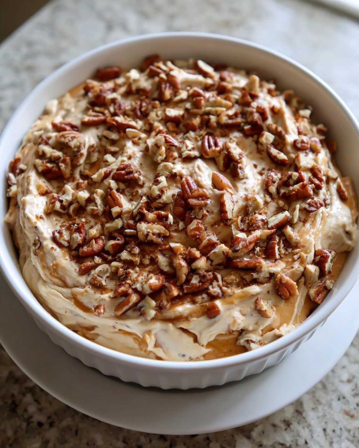 A white bowl filled with creamy, light brown pecan pie dip, generously topped with chopped pecans and caramel drizzle.