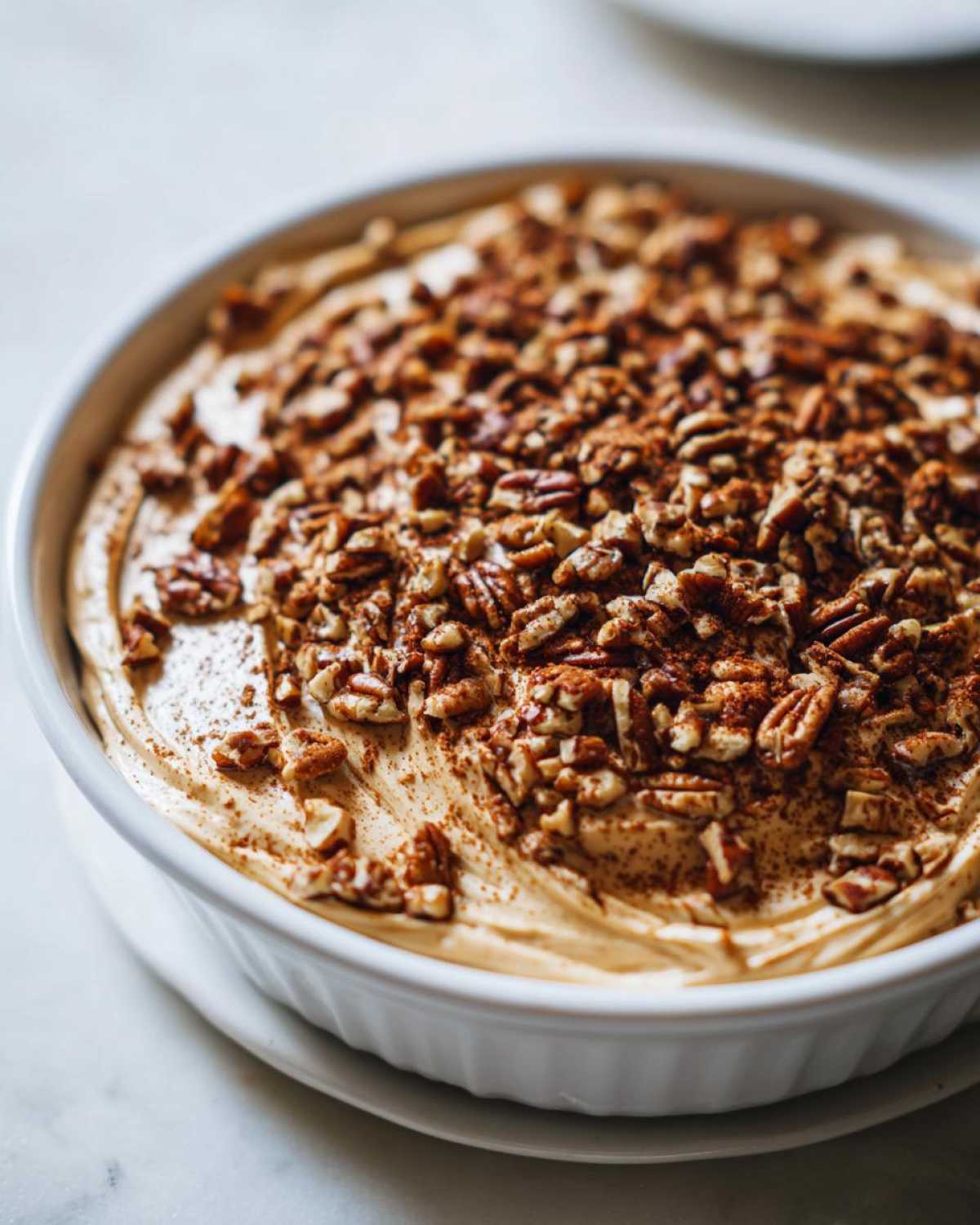 Close-up of a creamy, no-bake pecan pie dip in a white dish, generously topped with chopped pecans and cinnamon.
