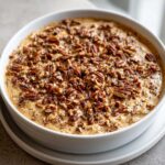 A creamy, caramel-colored no-bake pecan pie dip generously topped with chopped pecans in a white serving dish.