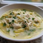 A close-up of a white bowl filled with steaming penicillin soup, featuring shredded chicken, orzo pasta, and parsley.