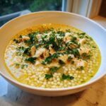 Close-up of a white bowl filled with penicillin soup, featuring small pasta pearls, shredded chicken, and topped with parsley and Parmesan.