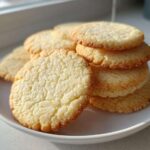 A stack of perfectly shaped, slightly crackled sugar cookie rounds resting on a white plate near a window.
