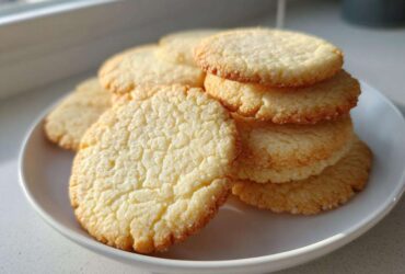 A stack of perfectly shaped, slightly crackled sugar cookie rounds resting on a white plate near a window.