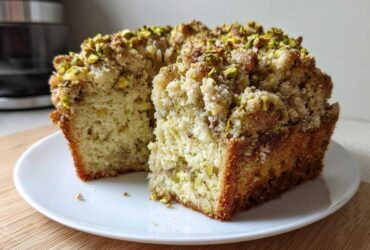 A cross-section of a moist pistachio coffee cake with a thick, sugary crumb topping sprinkled with chopped pistachios.