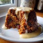 A rich, dark slice of traditional plum pudding covered in creamy custard sauce, served on a white plate.