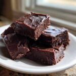 Close-up of rich, dark chocolate fudge squares stacked on a white plate, highlighting the smooth texture.