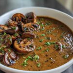 Close-up of a white bowl filled with rich brown mushroom sauce and whole sautéed mushrooms, garnished with parsley.
