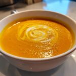 A close-up of creamy, bright orange roasted butternut squash soup, swirled with cream, served in a white bowl.