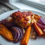 A piece of harissa chicken served on a white plate with roasted sweet potatoes and red onions.
