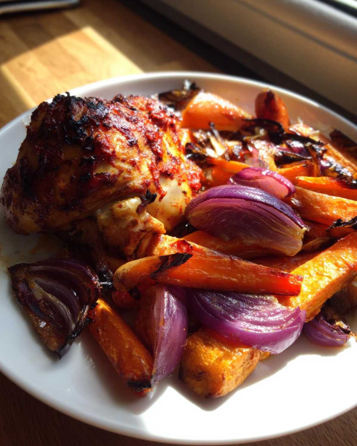 Close-up of a roasted chicken thigh coated in harissa spice served with roasted carrots and purple onions.