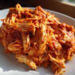 A close-up photo of shredded chicken tinga coated generously in a vibrant, rich red, smoky sauce, served on a white plate.
