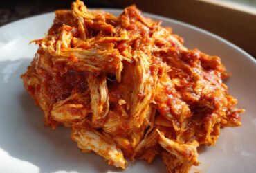 A close-up photo of shredded chicken tinga coated generously in a vibrant, rich red, smoky sauce, served on a white plate.