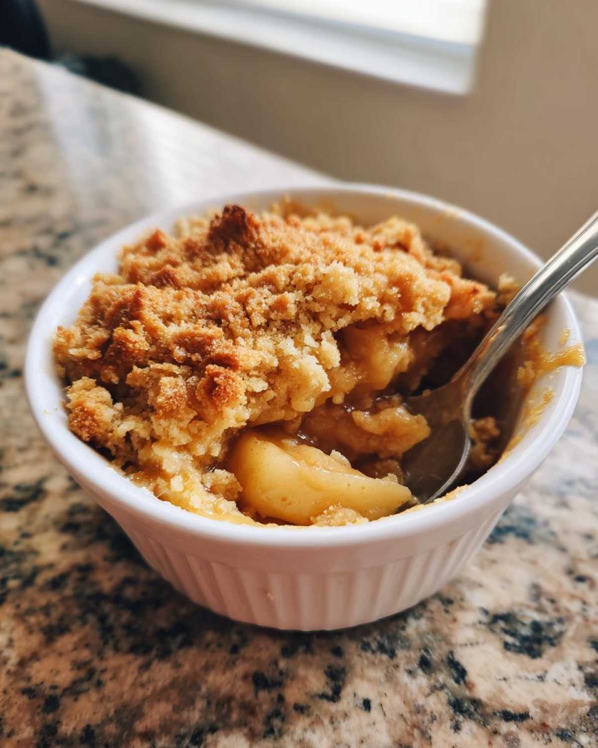 Close-up of a warm, individual serving of apple crumble with a golden brown topping in a white ramekin.