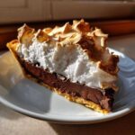 A close-up slice of rich chocolate meringue pie showing the dark filling and tall, toasted meringue.
