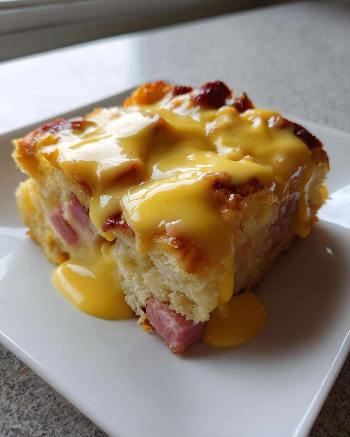 Close-up of a square slice of eggs benedict casserole topped generously with bright yellow hollandaise sauce.