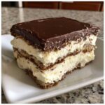 A close-up of a layered slice of eclair cake featuring graham crackers, creamy vanilla filling, and a rich chocolate topping.