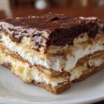 A close-up of a layered slice of eclair cake featuring graham crackers, white cream filling, and rich chocolate frosting.