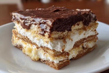 A close-up of a layered slice of eclair cake featuring graham crackers, white cream filling, and rich chocolate frosting.
