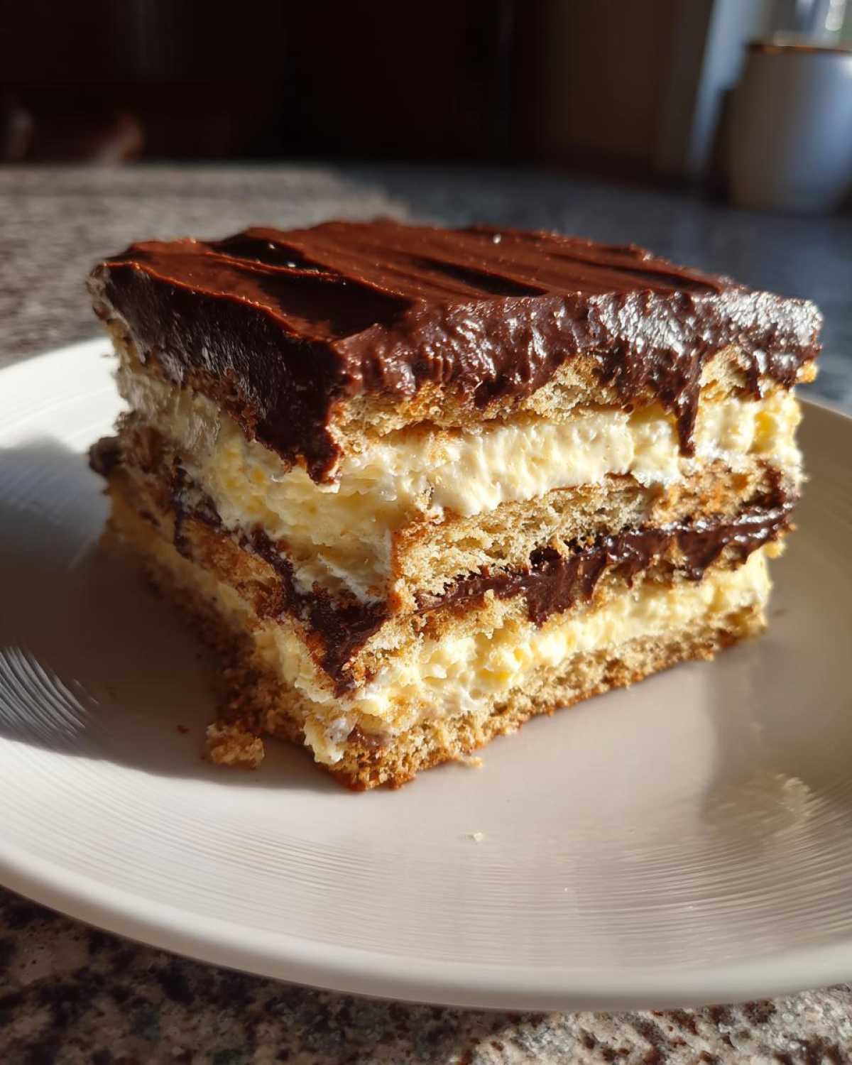 A thick slice of layered eclair cake featuring graham crackers, vanilla cream, and chocolate frosting, served on a white plate.