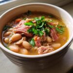 A close-up of a bowl of rich bean soup topped with shredded ham and fresh parsley.