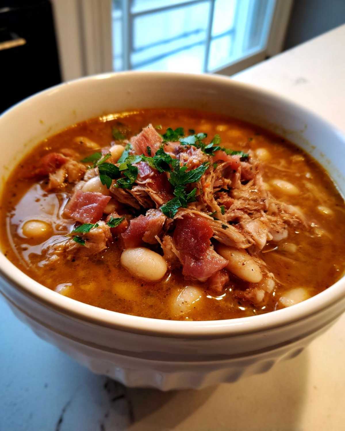 Close-up of a white bowl filled with rich, savory bean soup featuring white beans, shredded ham, and fresh parsley.