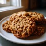 A close-up of several soft oatmeal cookies stacked on a white plate near a window.
