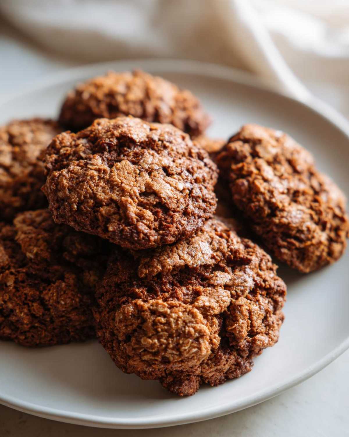 Close-up of several soft, dark brown oatmeal cookies stacked on a light-colored plate.