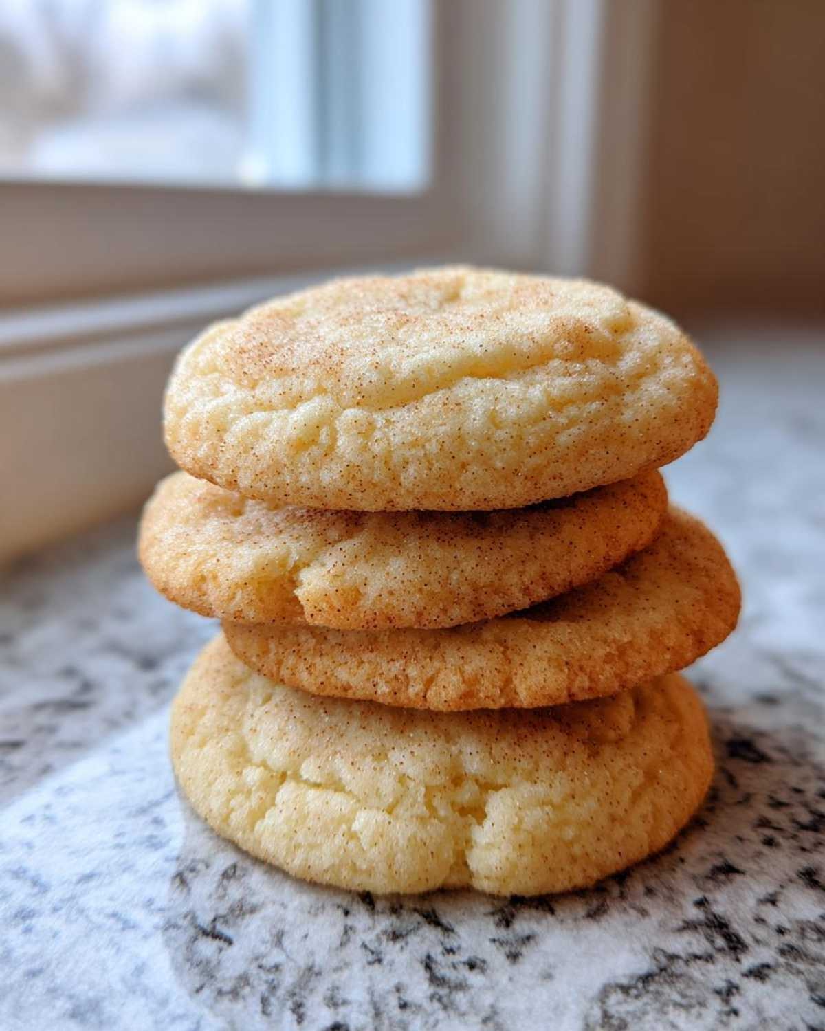 A stack of four soft sugar cookies coated in cinnamon sugar, sitting on a marble countertop.