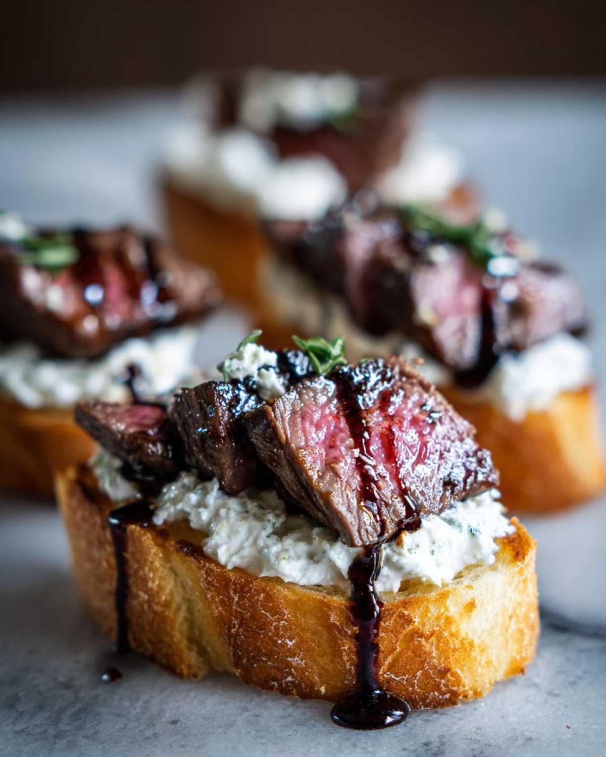 Close-up of one steak crostini topped with creamy cheese, sliced medium-rare steak, and a balsamic glaze drizzle.