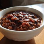 Close-up of a steaming white bowl filled with rich, thick black bean chili on a wooden surface.