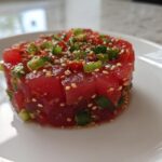 A close-up of a perfectly molded tower of vibrant red tuna tartare, topped with green onions and sesame seeds.
