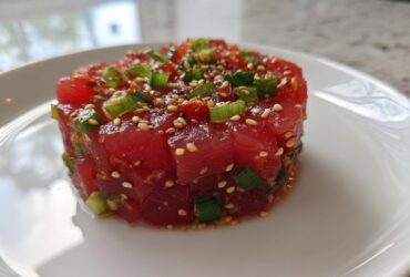 A close-up of a perfectly molded tower of vibrant red tuna tartare, topped with green onions and sesame seeds.