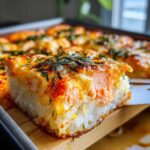 Close-up of a square slice of baked salmon sushi bake being lifted, showing layers of rice and creamy salmon topping.