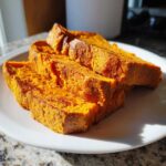 Three thick slices of vibrant orange sweet potato toast stacked slightly on a white plate, illuminated by sunlight.