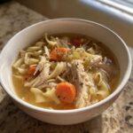 Close-up of a white bowl filled with homemade turkey noodle soup, featuring shredded turkey, egg noodles, and sliced carrots.