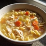 Close-up of a white bowl filled with steaming turkey noodle soup, featuring shredded turkey, egg noodles, and bright orange carrot slices.